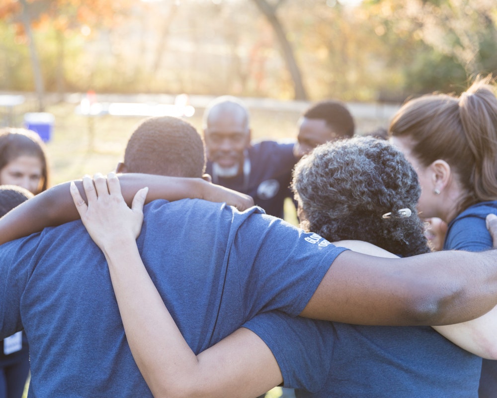 A diverse group of volunteers gathered into a huddle outside in a park.