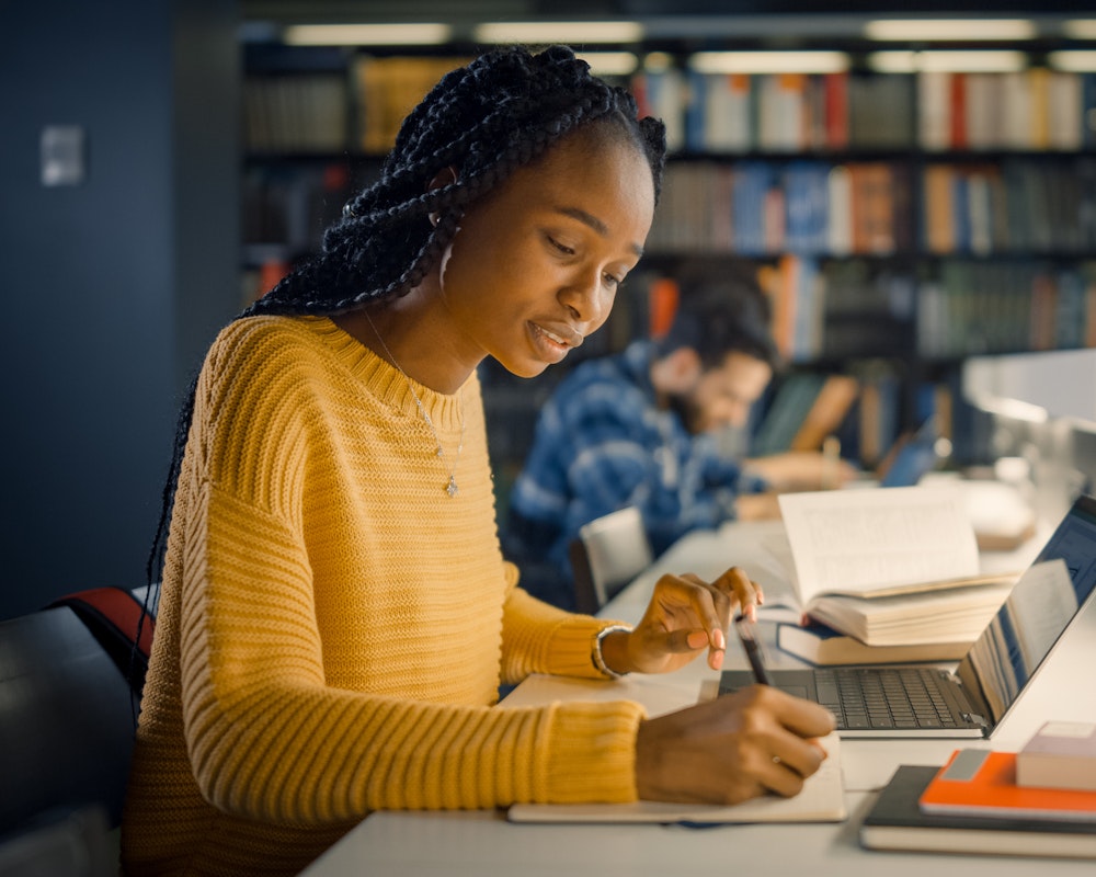 A Black student in a yellow sweater studies in a library